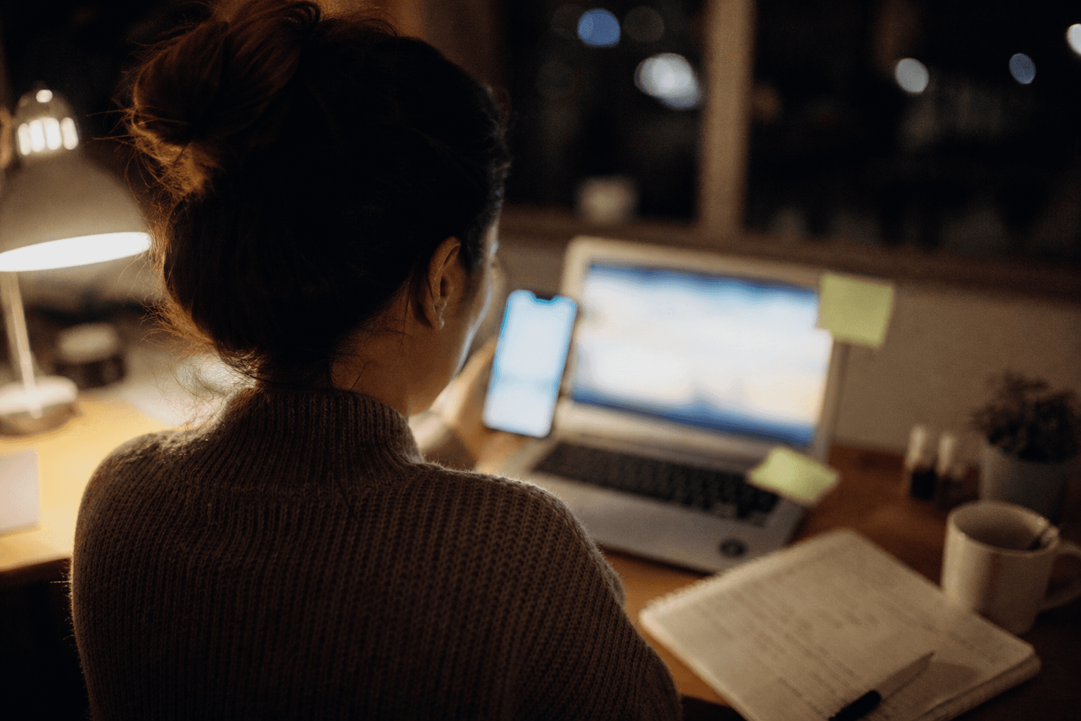 A homeowner researching contractors at night, phone in hand with a laptop open beside her