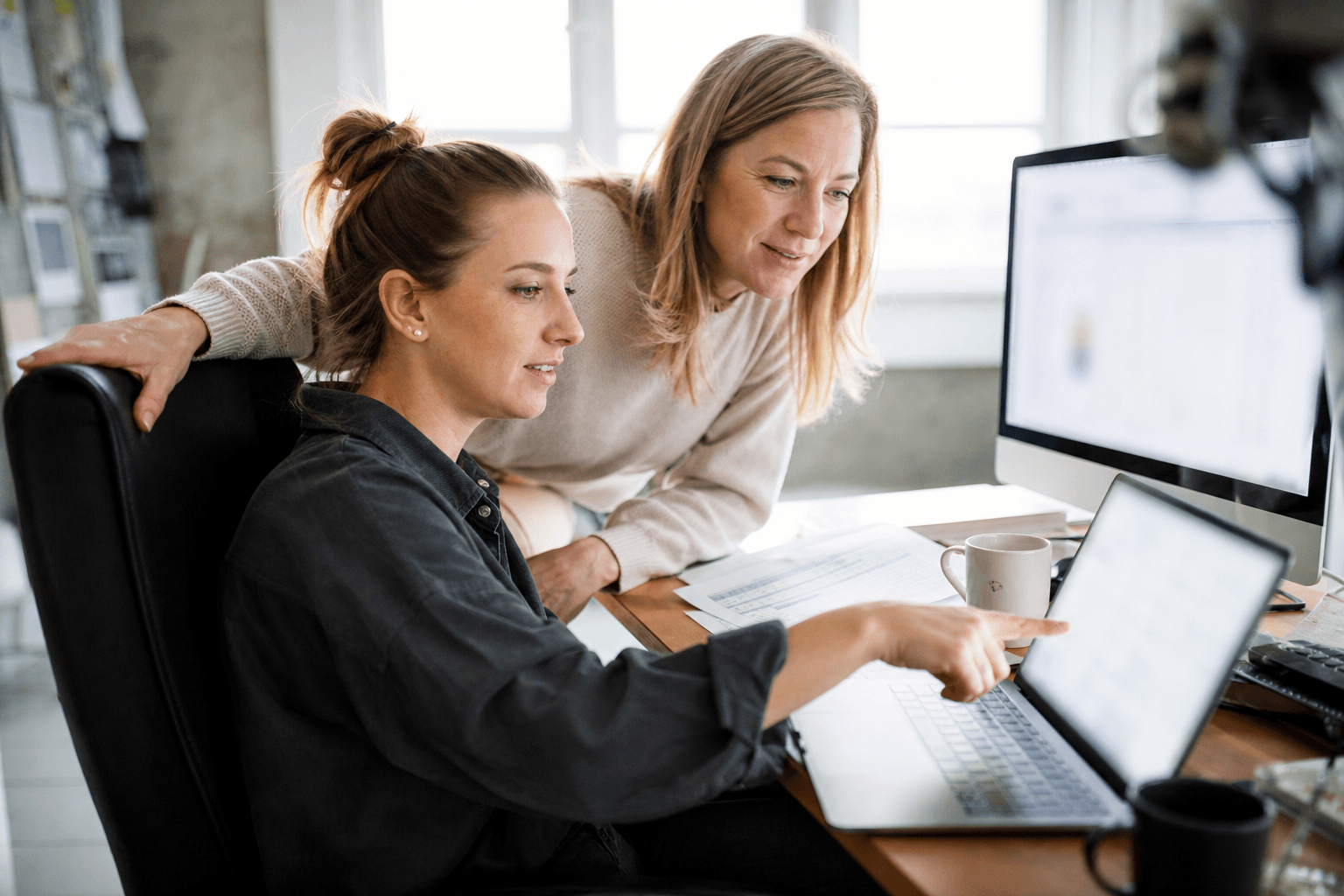 Two colleagues comparing software dashboards side by side on a laptop and a desktop monitor